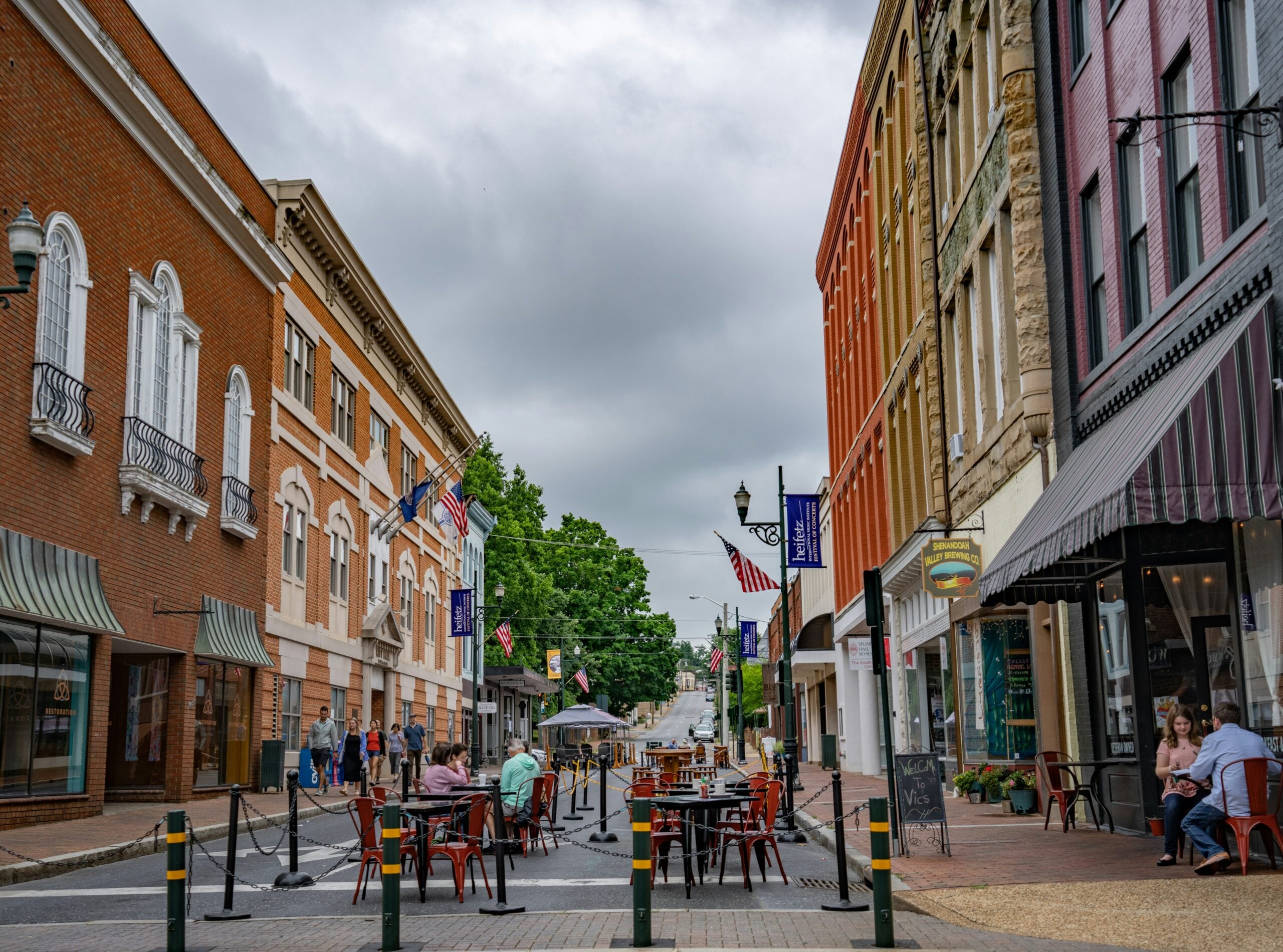 People dine at outdoor tables set up in the middle of a small town street lined with historic brick buildings, colorful storefronts, and American flags on a cloudy day.