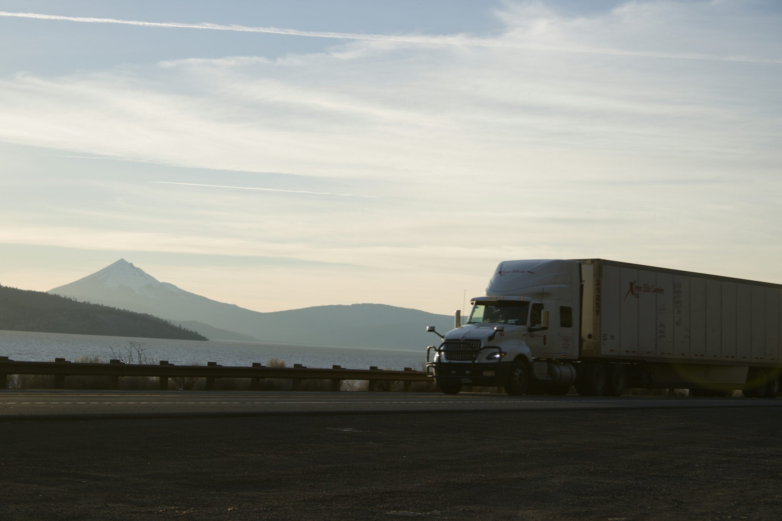 A semi-truck drives along a highway near a body of water at sunset, with mountains and a snow-capped peak visible in the background.