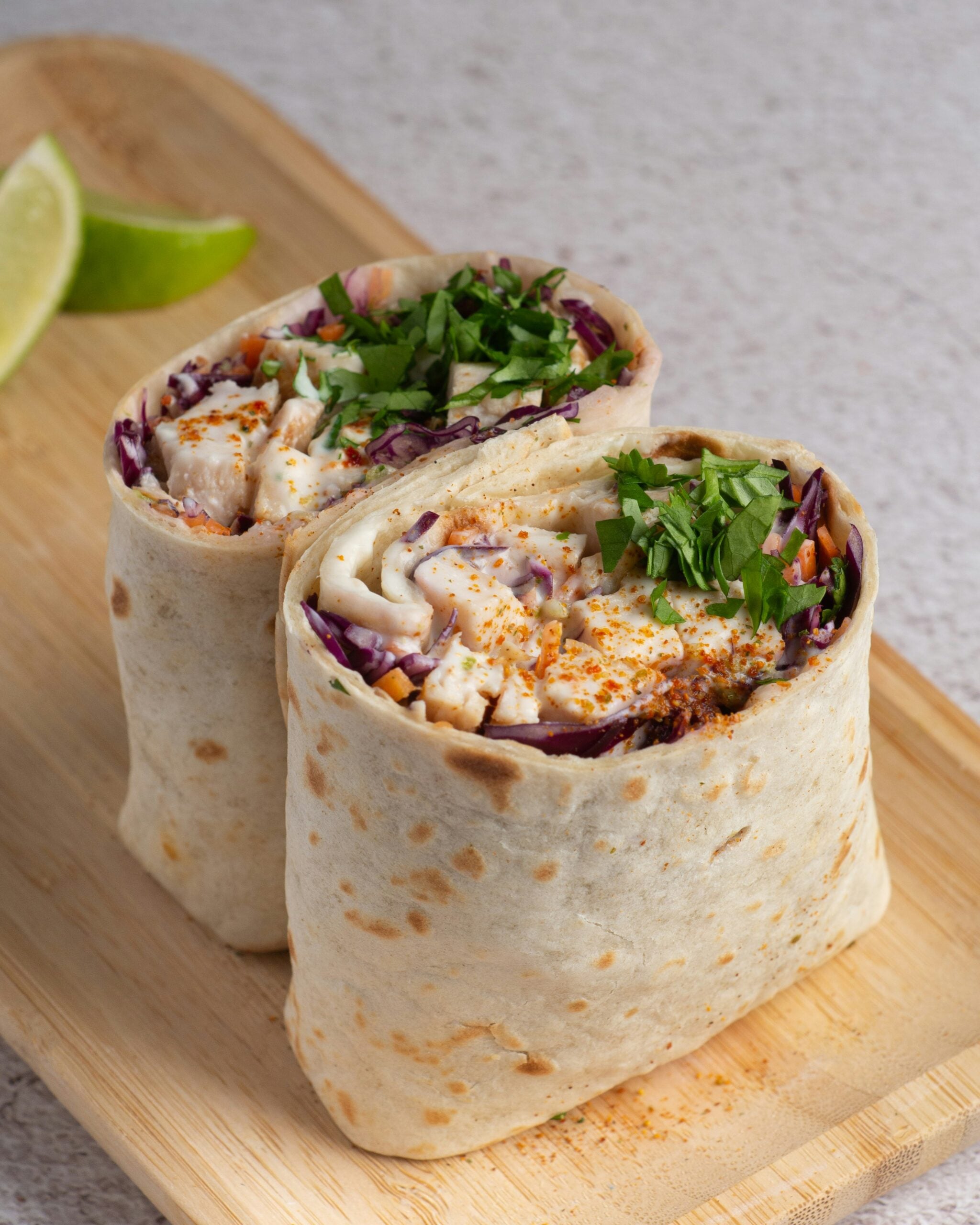 A close-up of a wrap cut in half, filled with grilled chicken, shredded red cabbage, chopped cilantro, and spices, served on a wooden board with lime wedges in the background.