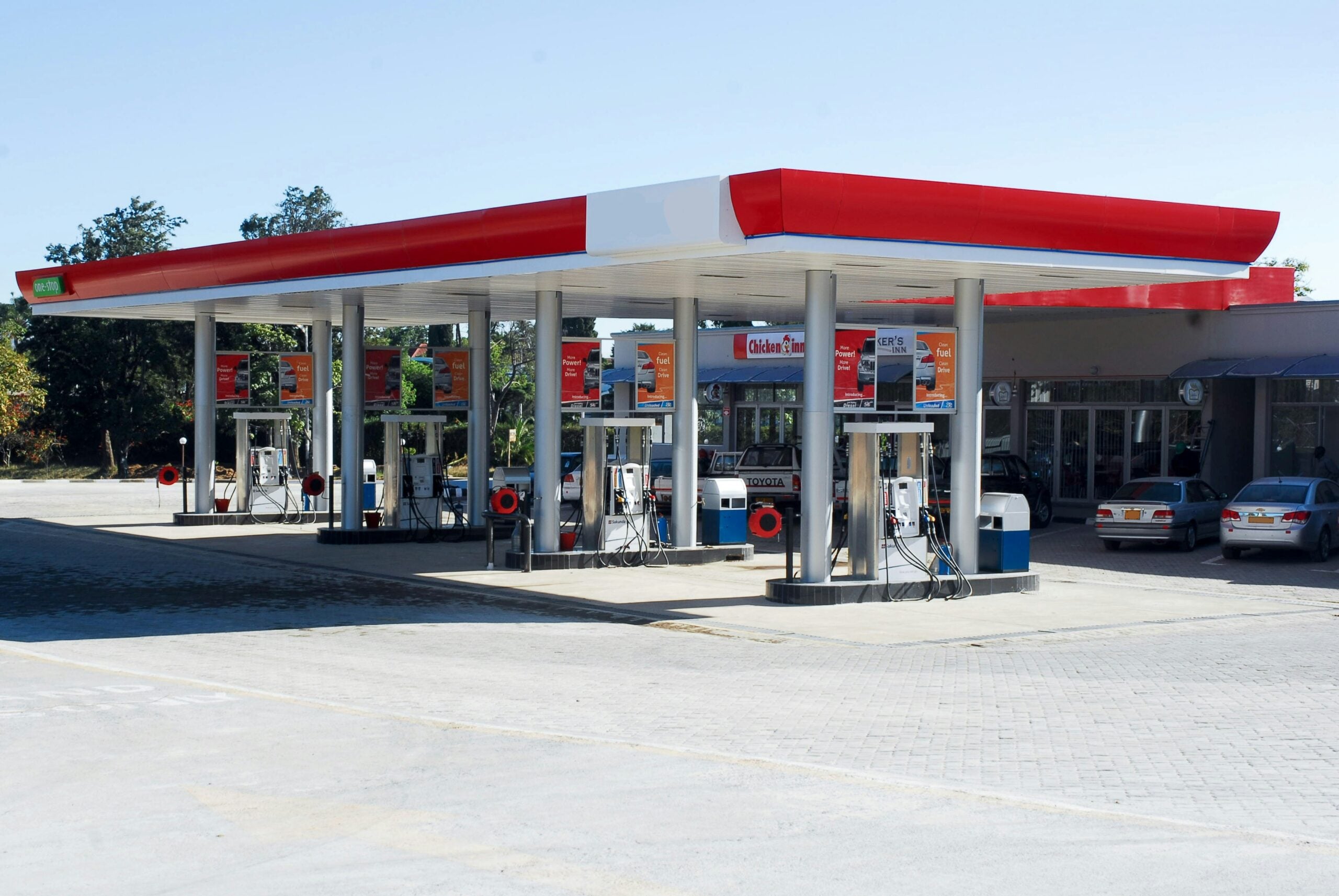 A modern gas station with multiple fuel pumps and a red and white canopy, set on a paved lot with a few parked cars and a small building in the background on a clear, sunny day.