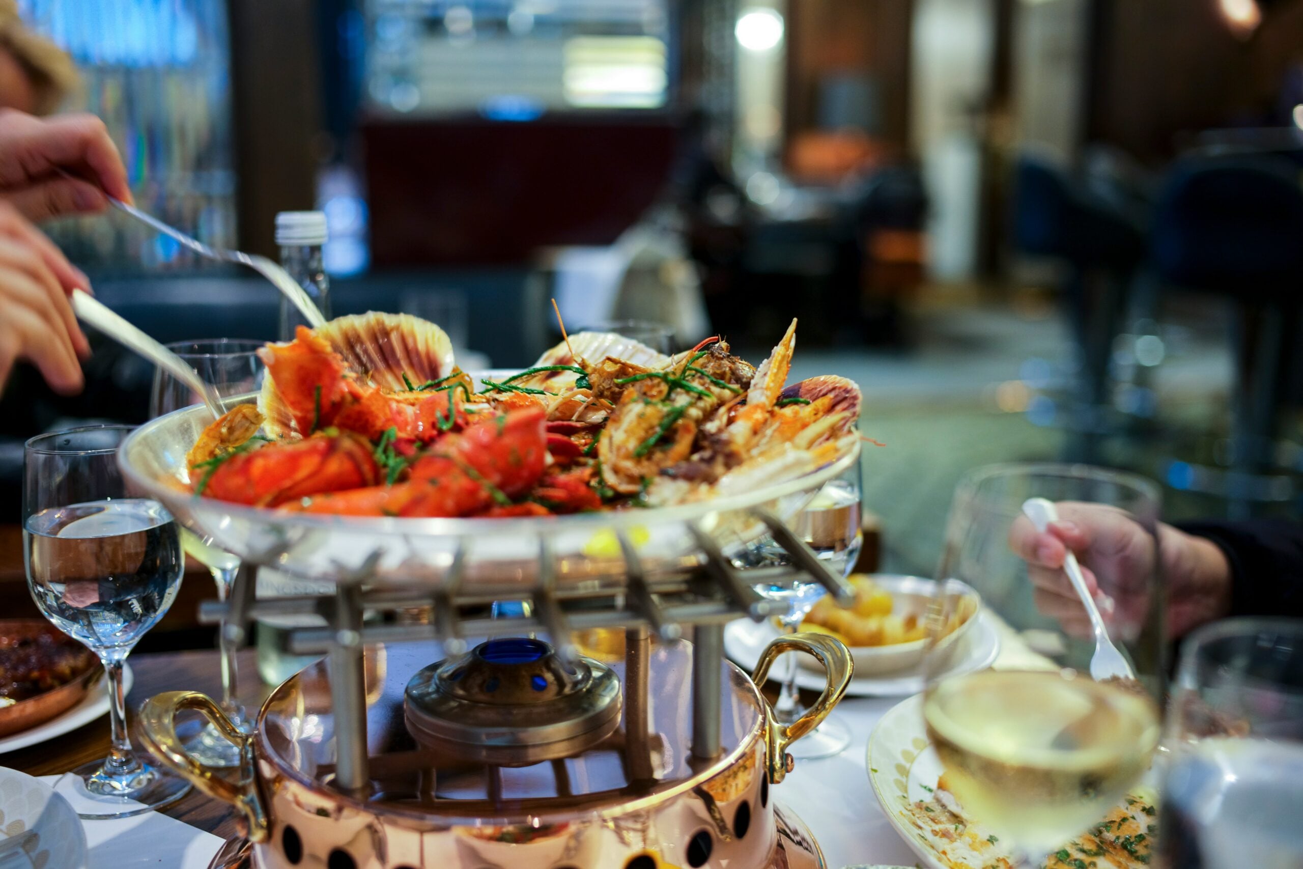 A large seafood platter with lobster, shellfish, and garnish is served on a raised dish at a restaurant table, surrounded by glasses of white wine and people dining.