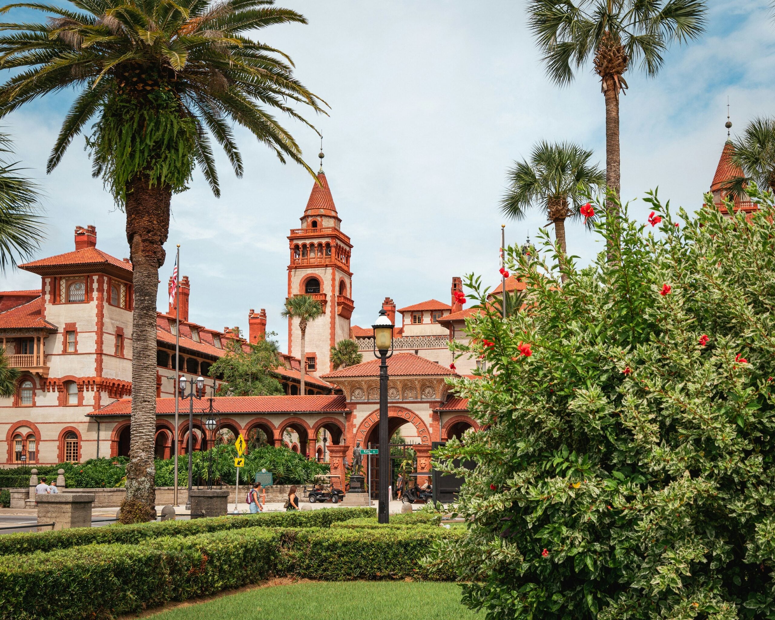 A historic red-roofed building with towers and arched windows surrounded by palm trees, manicured hedges, and flowering bushes under a partly cloudy sky.