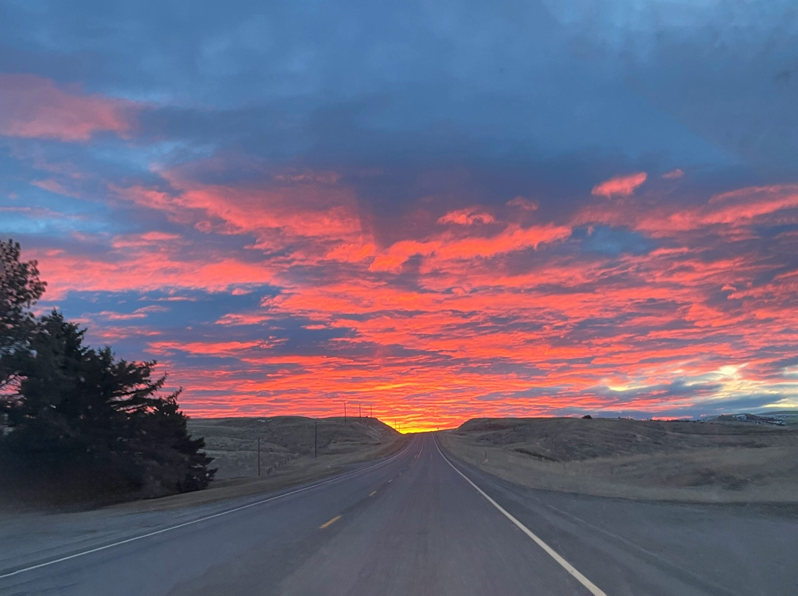 A paved road stretches into the distance under a dramatic sunset sky filled with vibrant orange, pink, and blue clouds. The landscape is mostly open with a few trees and gentle hills on either side.