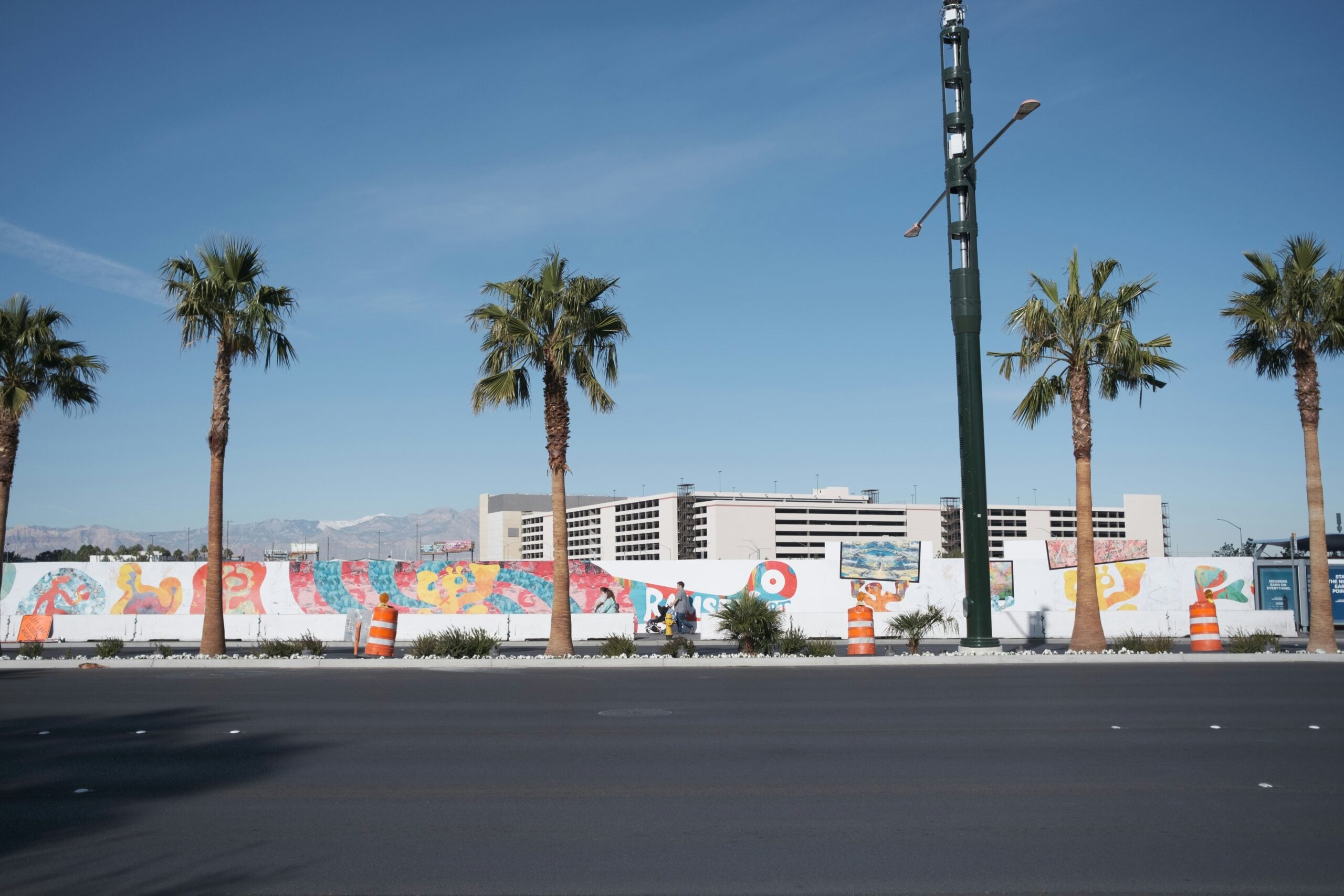 A city street with palm trees, orange construction barrels, and a colorful mural on a wall. A large building is visible in the background under a clear blue sky.