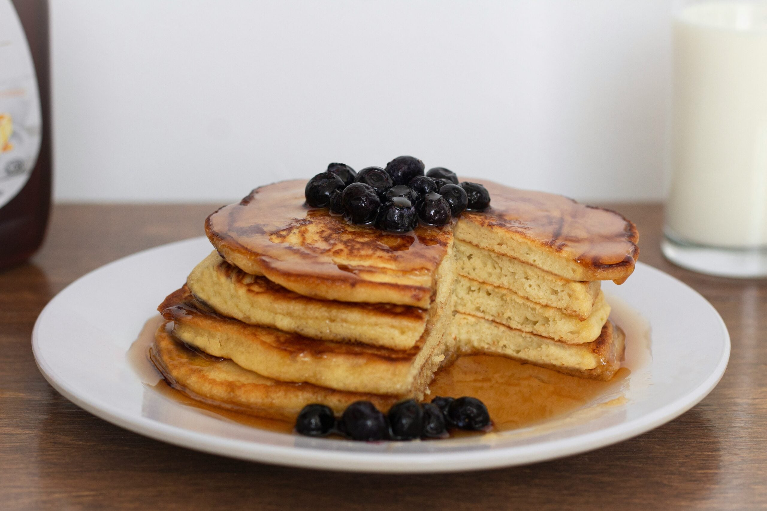 A stack of fluffy pancakes topped with blueberries and syrup sits on a white plate. A glass of milk and a syrup bottle are in the background on a wooden surface.