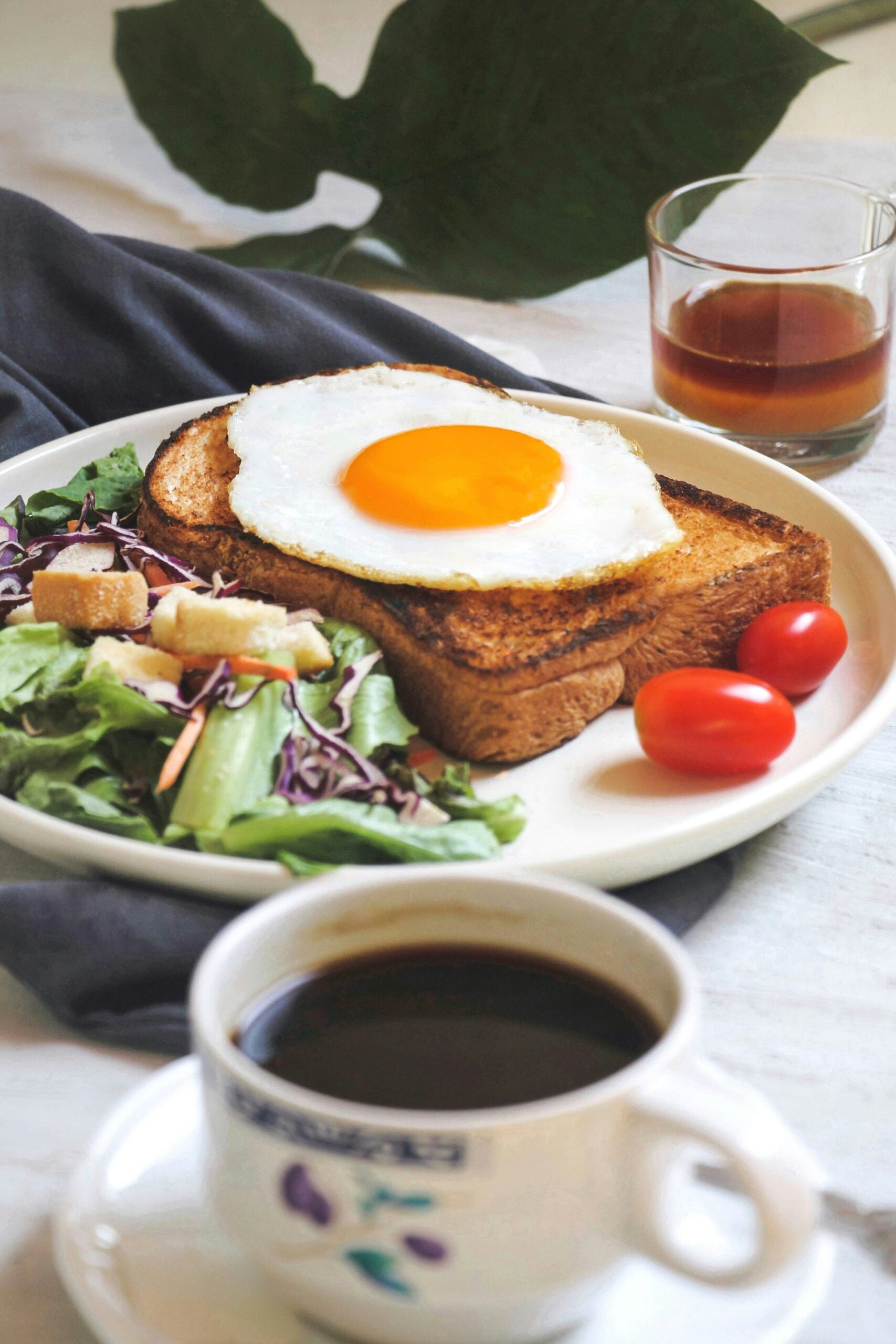 A breakfast plate with two slices of toast topped with a sunny-side-up egg, cherry tomatoes, green salad with croutons, a glass of juice, and a cup of black coffee. A dark napkin and a large green leaf are in the background.
