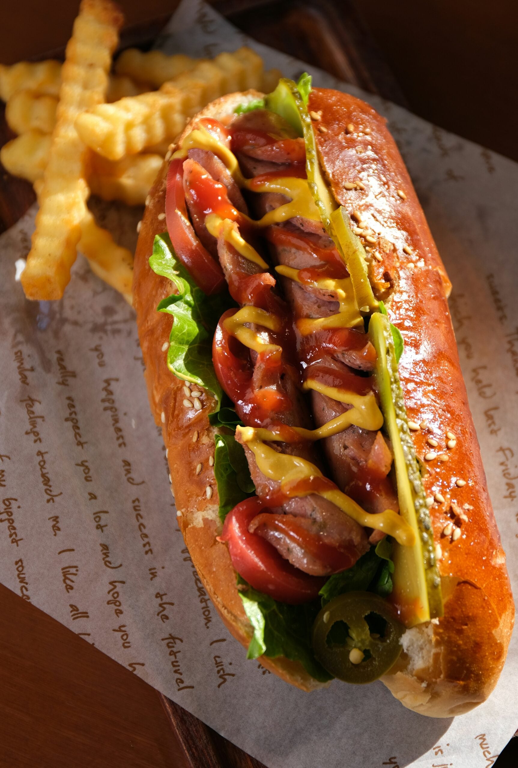 A hot dog topped with sliced tomatoes, lettuce, pickles, jalapeños, ketchup, and mustard, served on a sesame bun with a side of crinkle-cut fries on parchment paper.