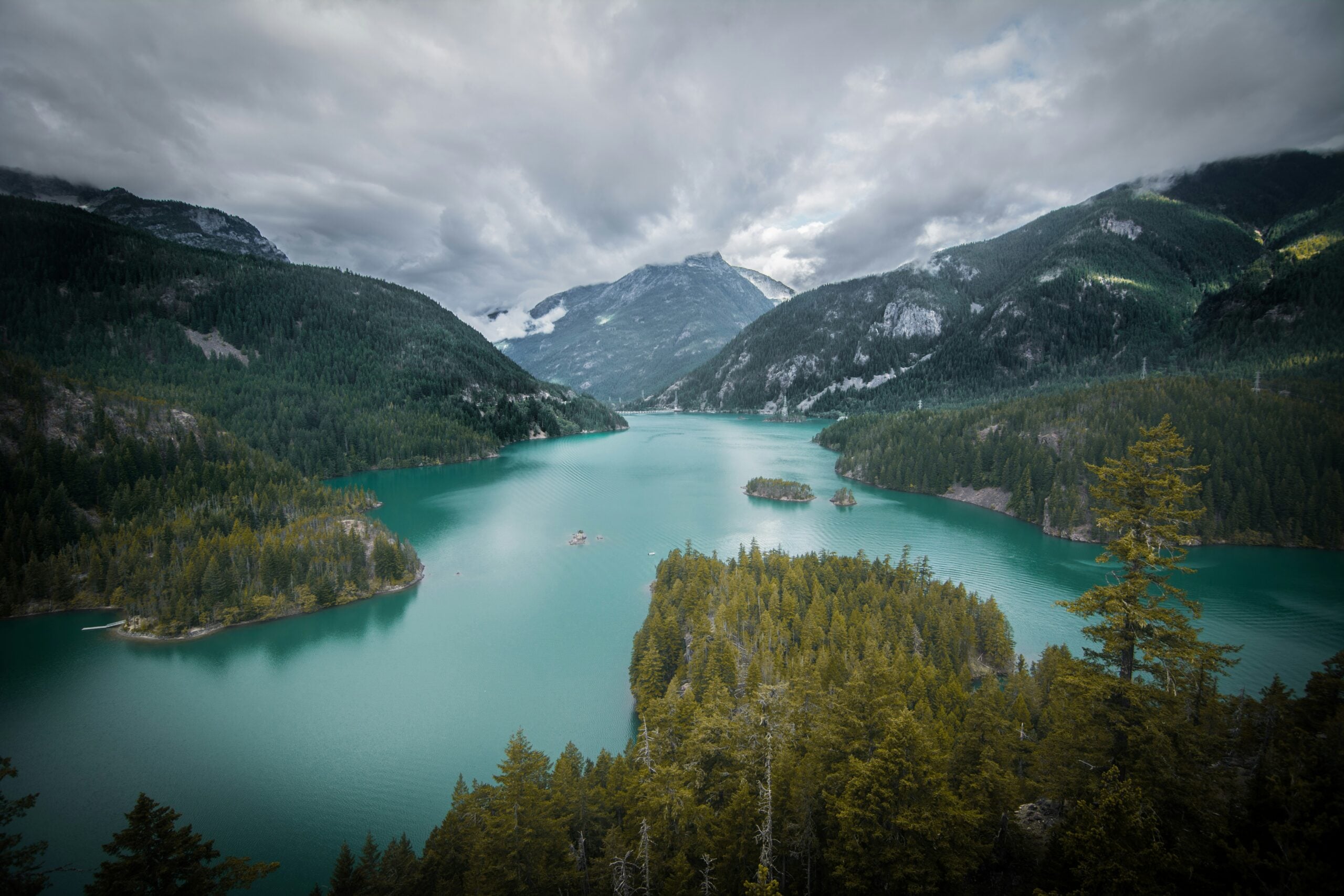 A scenic view of a turquoise lake surrounded by dense evergreen forests and mountains under a cloudy sky, with small islands visible in the water.