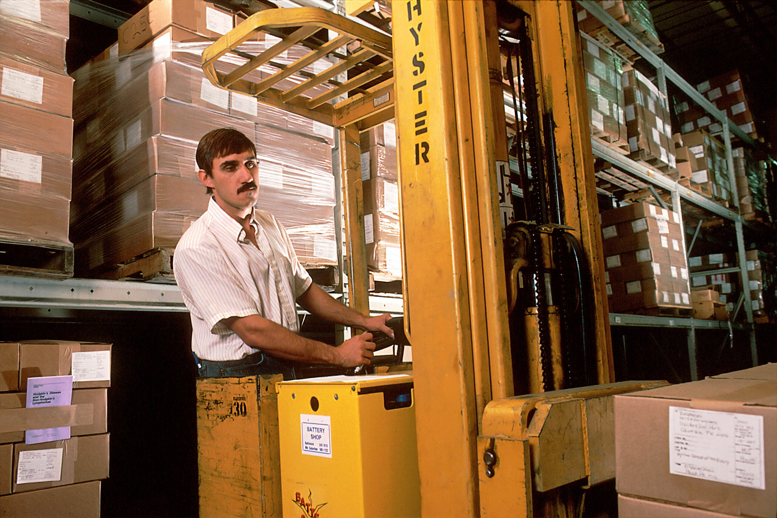 A man operating a yellow forklift in a warehouse filled with stacked boxes and shelves, wearing a striped shirt and surrounded by packages.