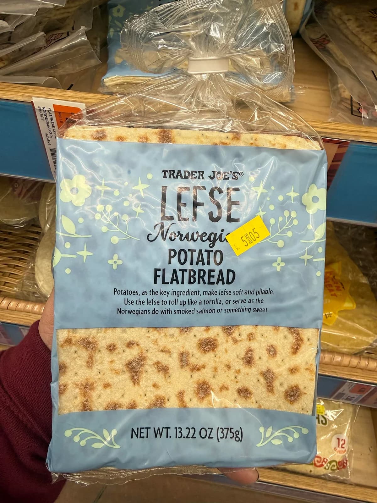 A hand holds a package of Trader Joe's Lefse Norwegian Potato Flatbread, 13.22 oz (375g), with a yellow price sticker showing $5.49. The packaging features blue floral designs.