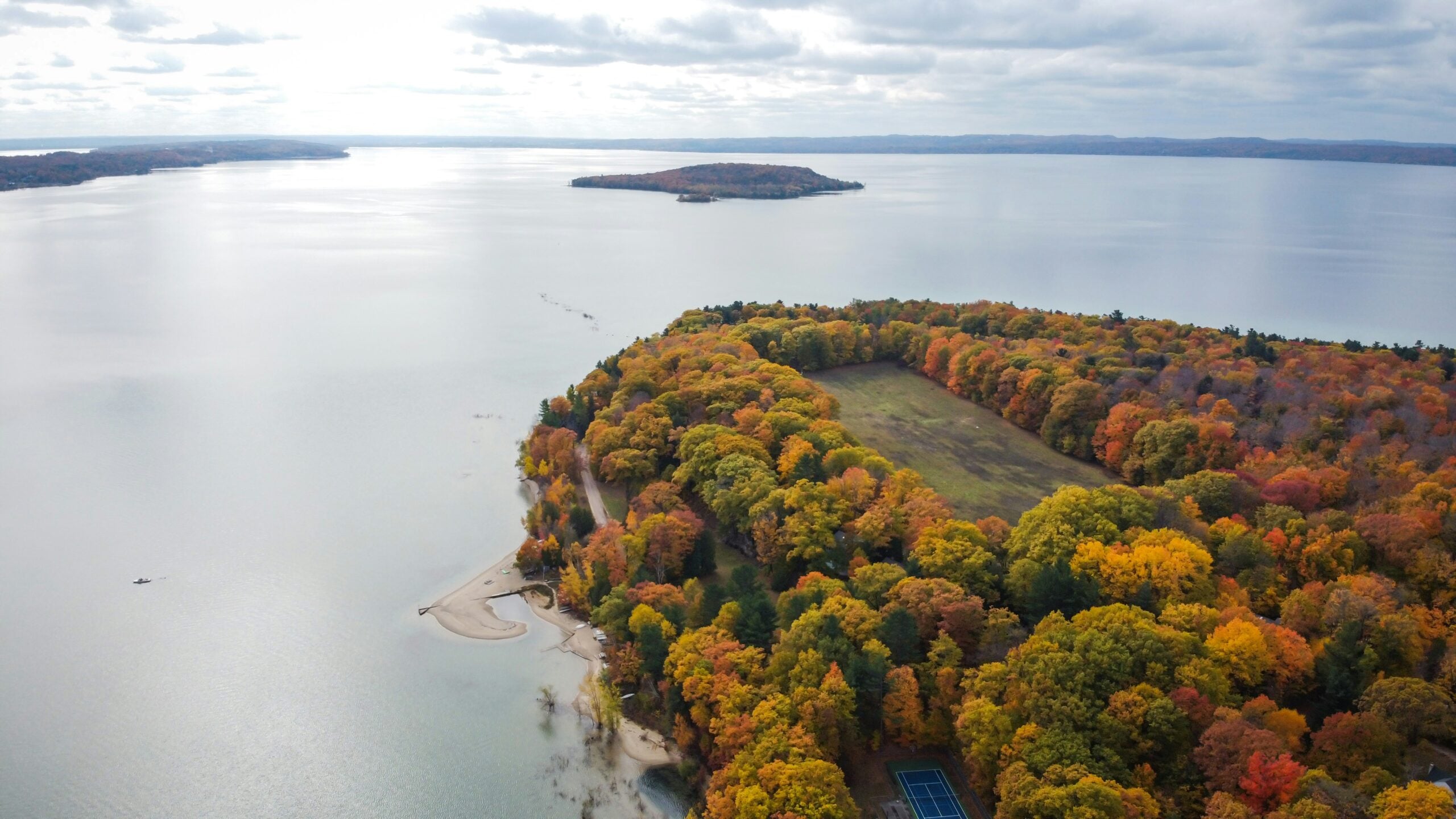 Aerial view of a peninsula covered in autumn trees beside a calm lake, with a small sandy beach and tennis court visible, under a cloudy sky. An island can be seen in the distance.
