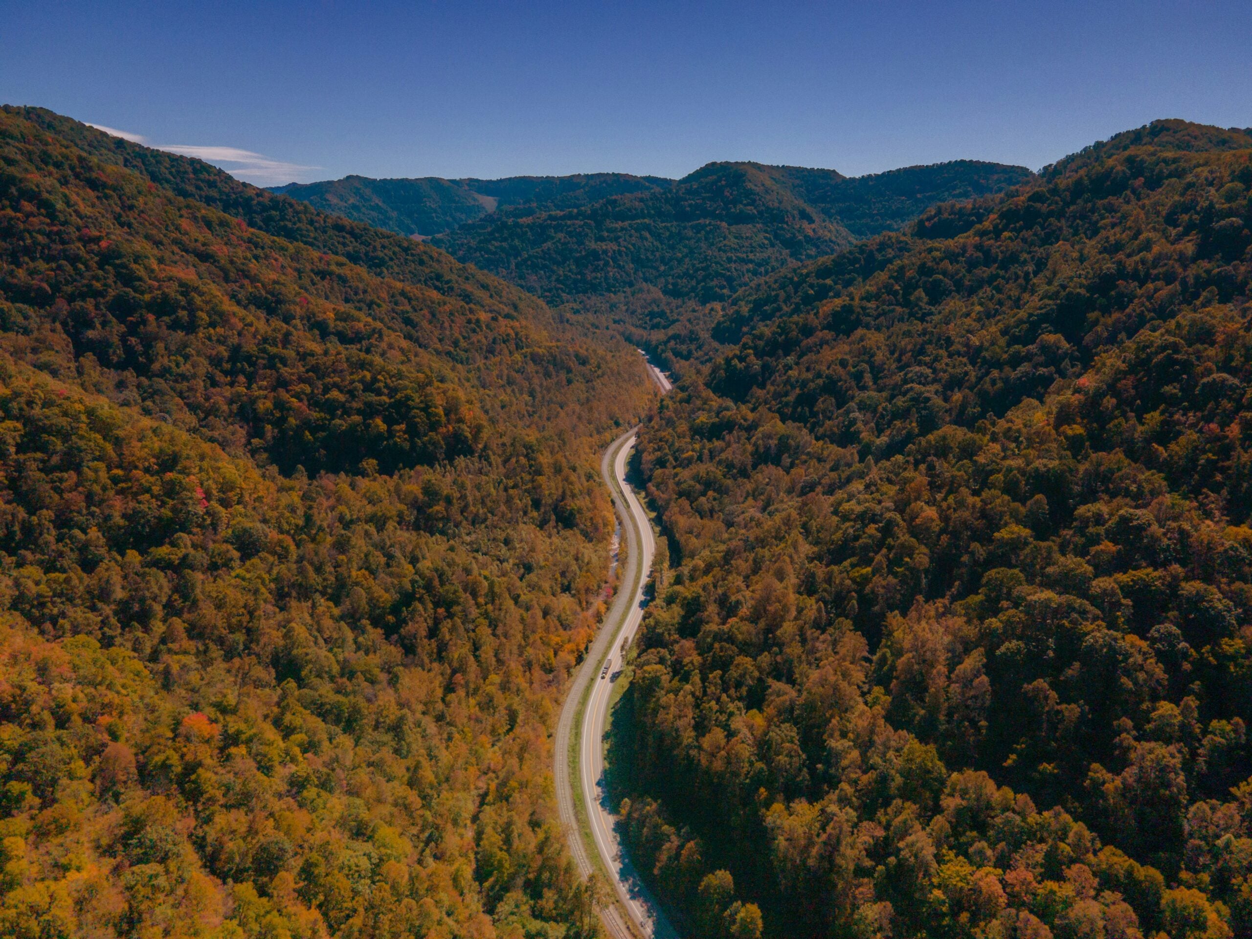 A winding road cuts through a dense, forested valley surrounded by rolling green hills and mountains under a clear blue sky.