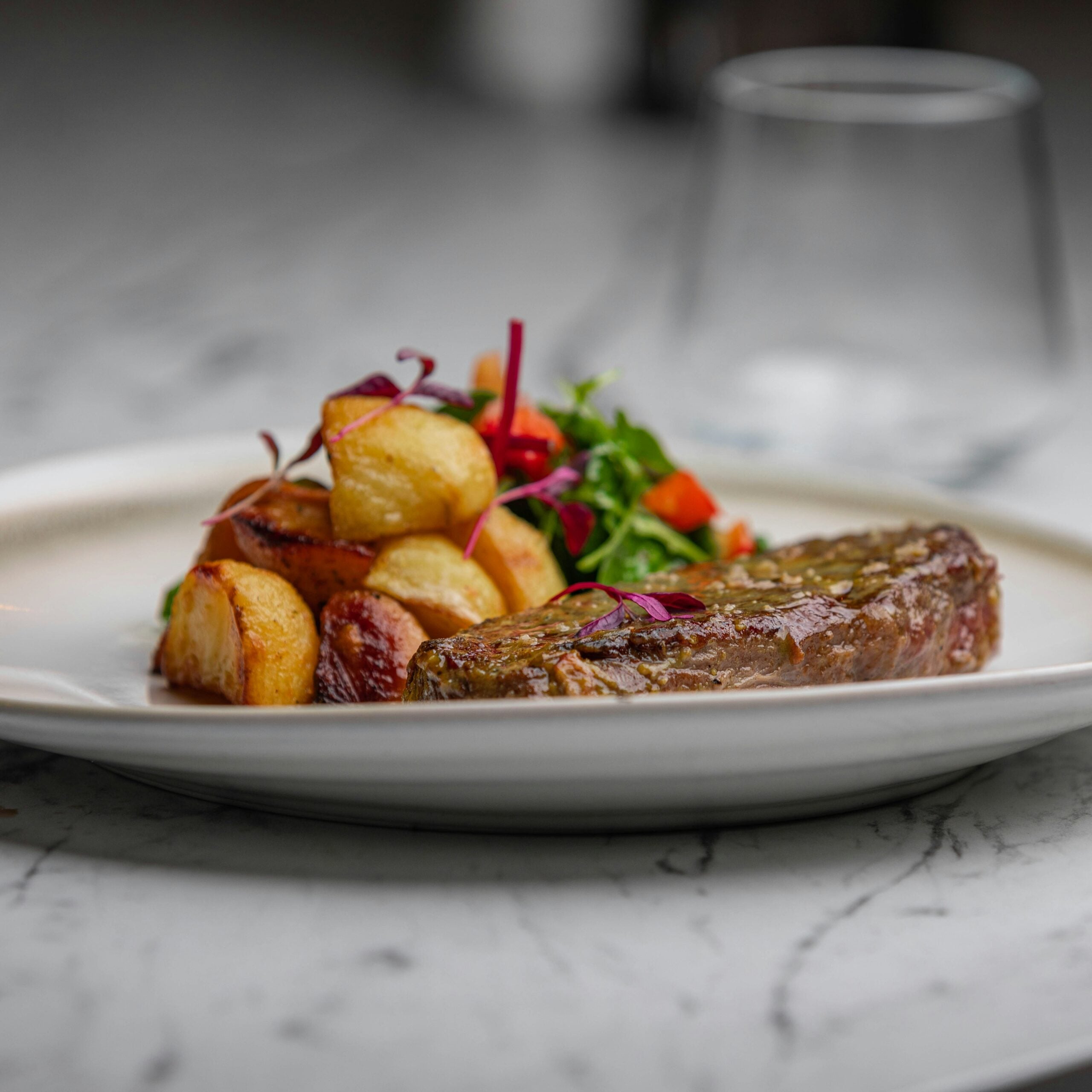 A plated meal featuring a grilled steak, golden roasted potatoes, and a colorful garnish of greens and vegetables, set on a marble table with a blurred glass in the background.
