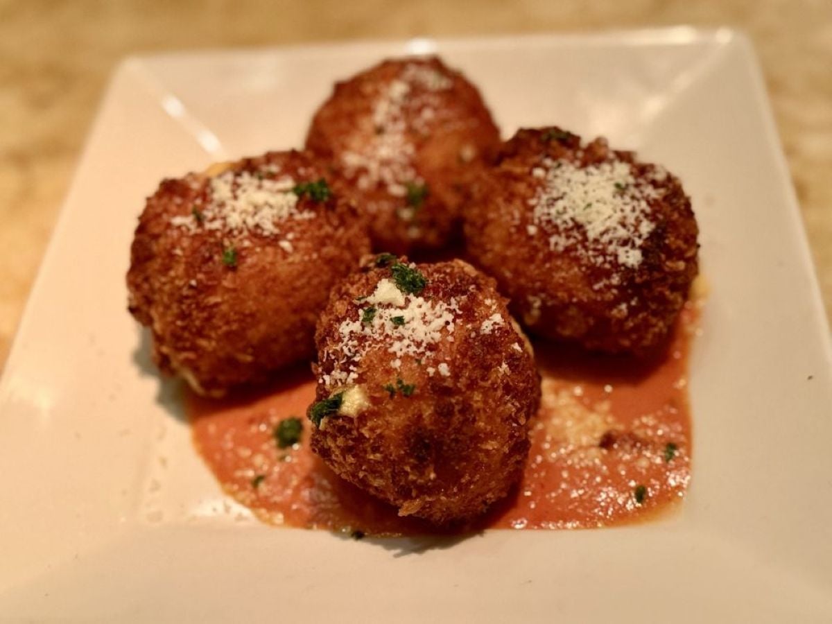 Four golden-brown, breaded arancini balls topped with grated cheese and herbs, served on a square white plate over a layer of tomato sauce.