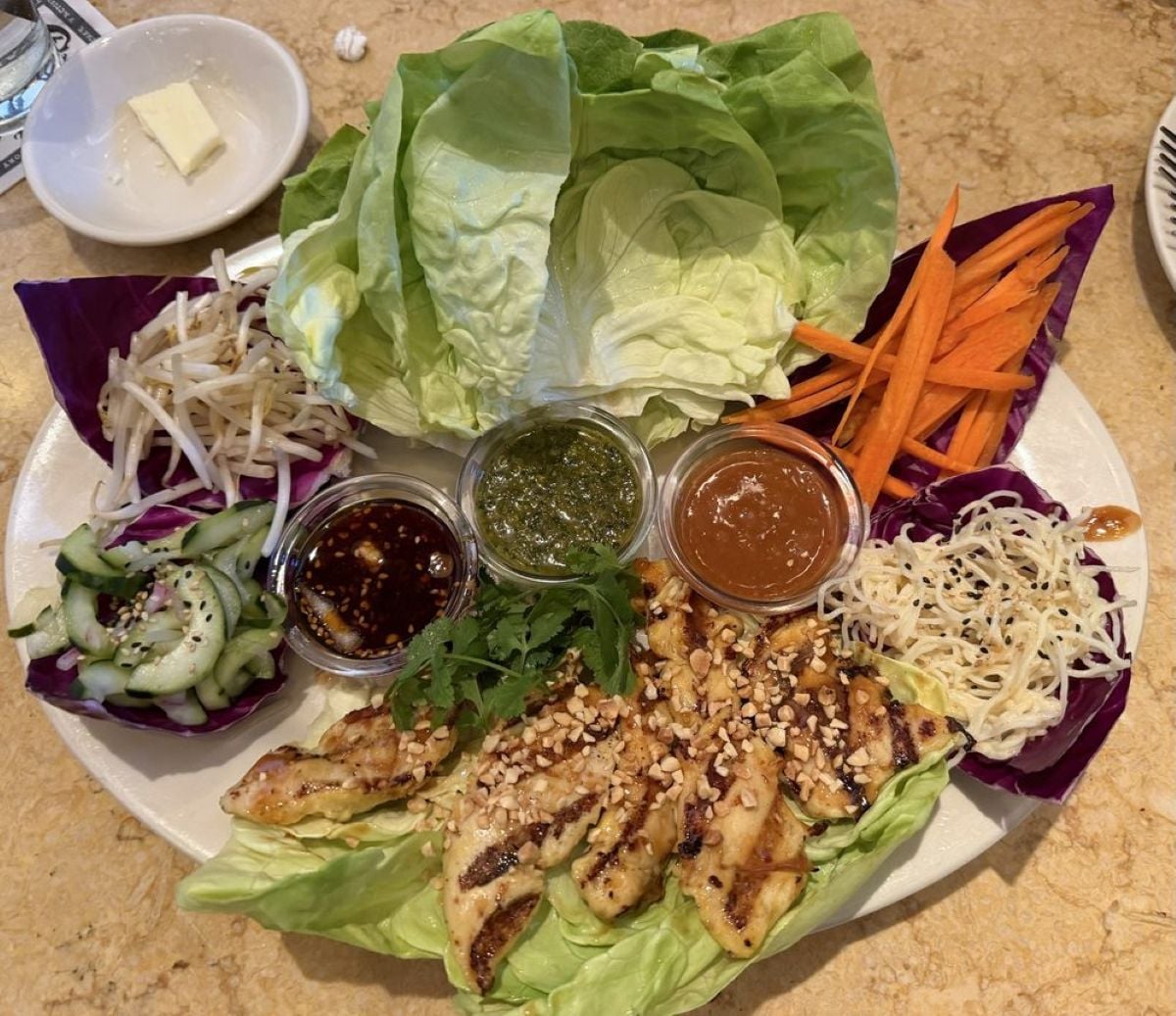 A plate with grilled chicken, lettuce leaves, shredded carrots, red cabbage, bean sprouts, cucumber salad, noodles, and three dipping sauces, garnished with herbs and chopped peanuts. A pat of butter is in the background.