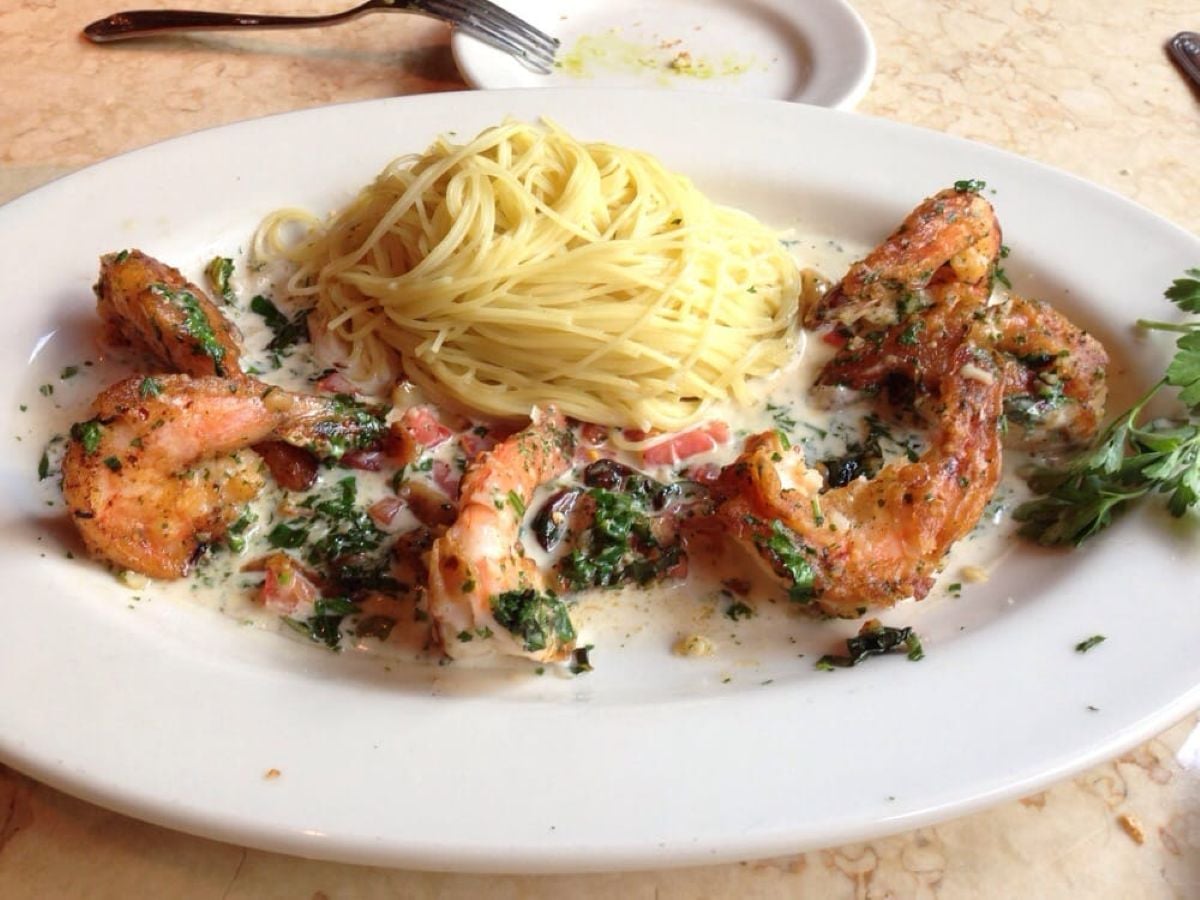 A white plate with a serving of pasta next to five large shrimp in a creamy herb sauce, garnished with chopped tomatoes and greens, with a sprig of parsley on the side. A fork and small plate are in the background.