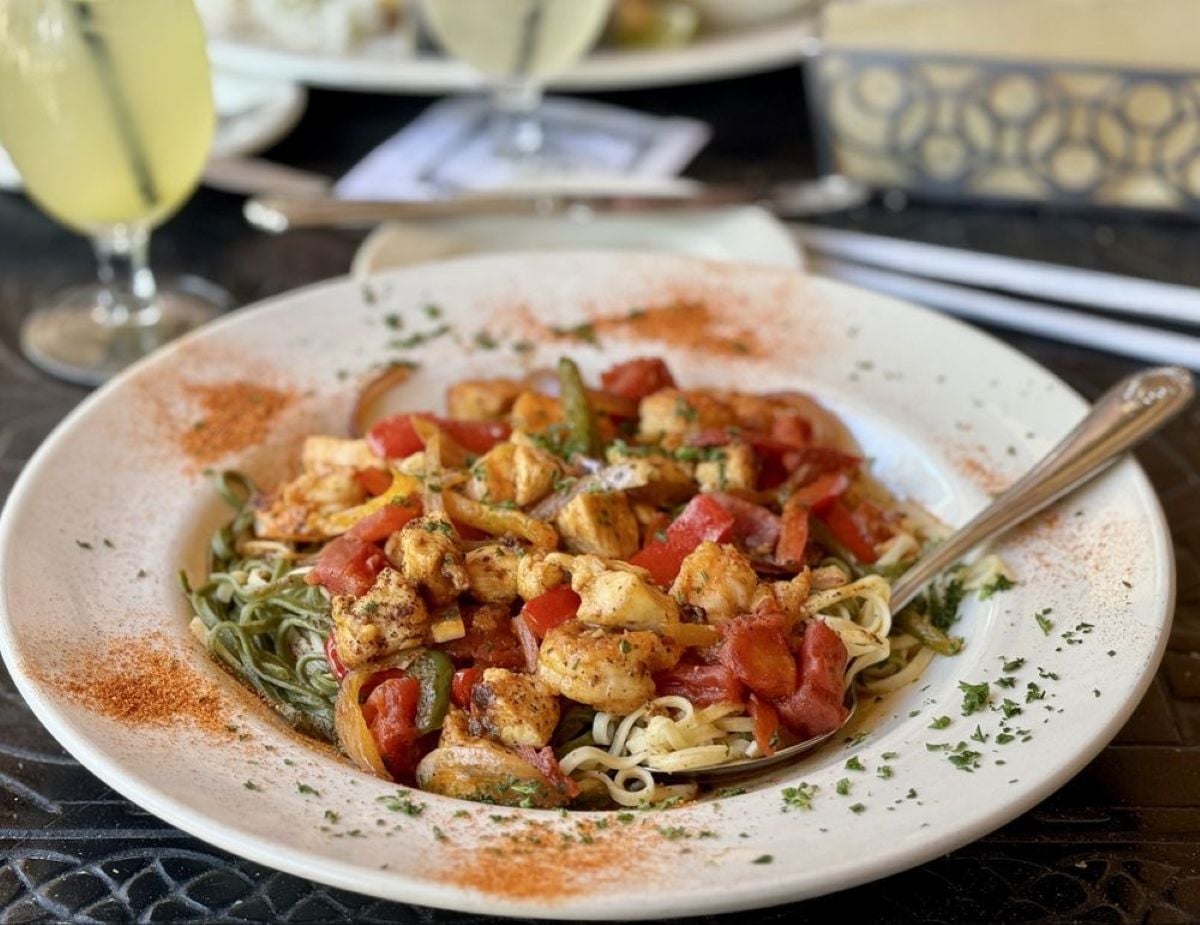 A plate of pasta topped with grilled shrimp, diced tomatoes, herbs, and spices, with a fork resting on the dish. A glass of yellow beverage and another plate are visible in the blurred background.
