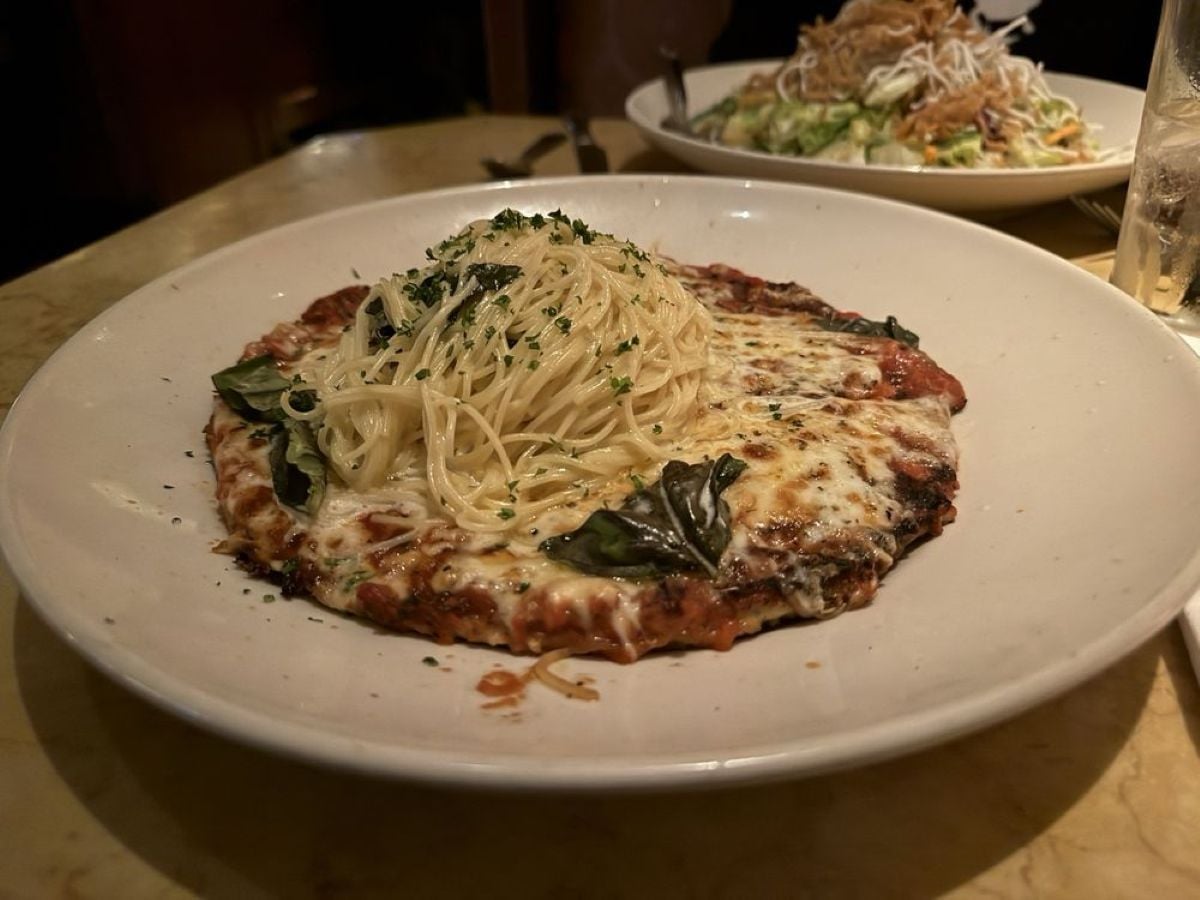 A plate with a cheesy, baked pizza topped with fresh basil, and a serving of plain spaghetti pasta placed on top. Another plate of salad is partially visible in the background.