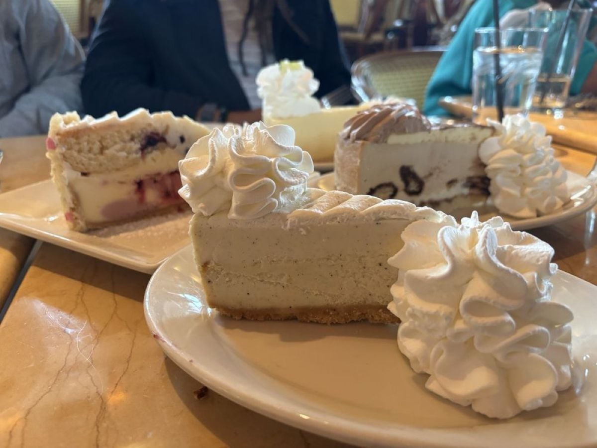 Three slices of cheesecake topped with whipped cream are on white plates, with people and glasses of water in the background at a restaurant.