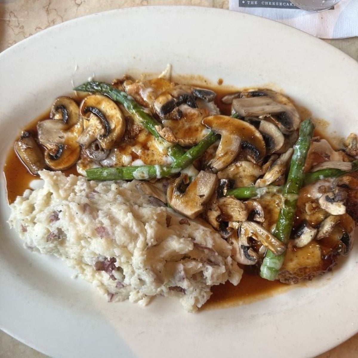 A white plate with mashed potatoes, grilled chicken breast topped with sautéed mushrooms, asparagus, and brown sauce.
