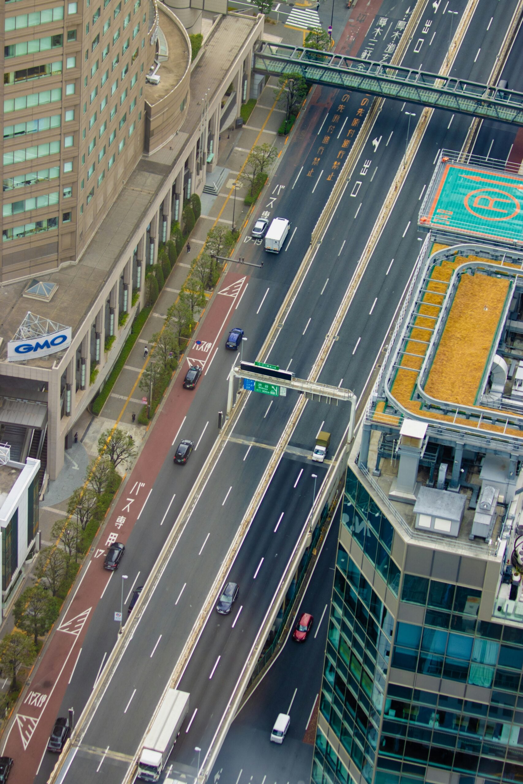 Aerial view of a city street with cars and trucks on multiple lanes, a pedestrian bridge, and adjacent high-rise buildings, including one with a rooftop helipad. Trees line the sidewalk.