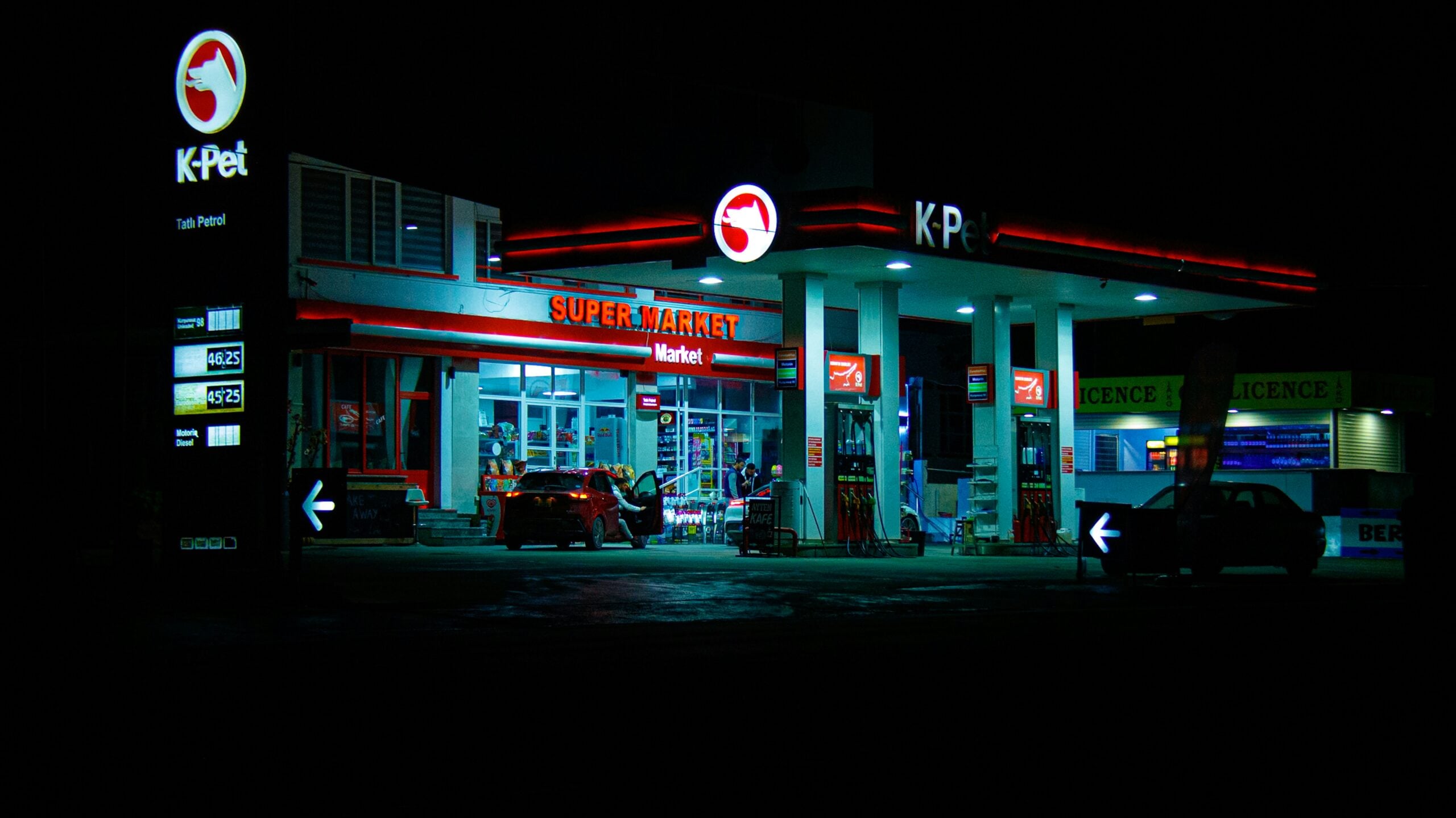 A brightly lit K-Pet gas station and supermarket at night, with cars parked by the fuel pumps and illuminated signs displaying fuel prices. The surrounding area is dark.