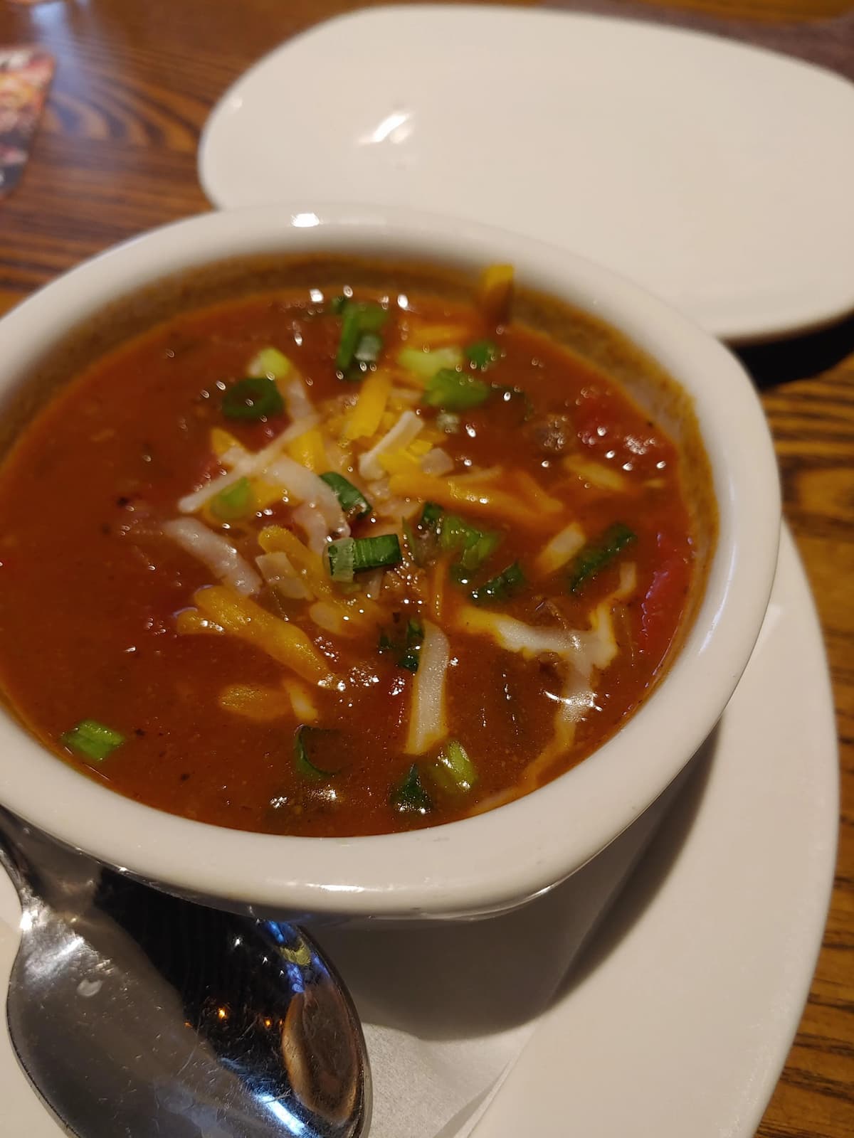 A bowl of chili topped with shredded cheese and chopped green onions sits on a white plate with a spoon beside it, ready to be served.