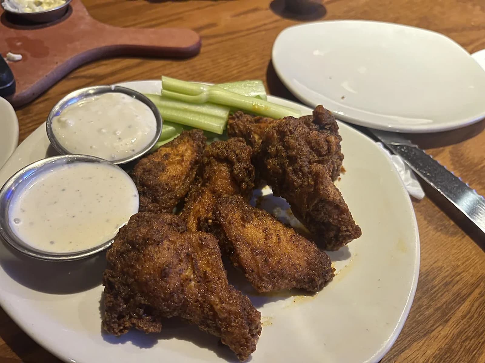A plate of crispy chicken wings served with celery sticks and two small cups of ranch or blue cheese dipping sauce on a white plate, with a knife and an empty plate nearby.