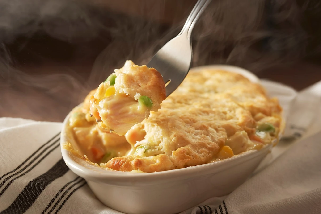 A fork lifts a steaming bite of creamy chicken pot pie topped with golden biscuit crust from a small white baking dish, showing pieces of chicken, peas, carrots, and corn inside.
