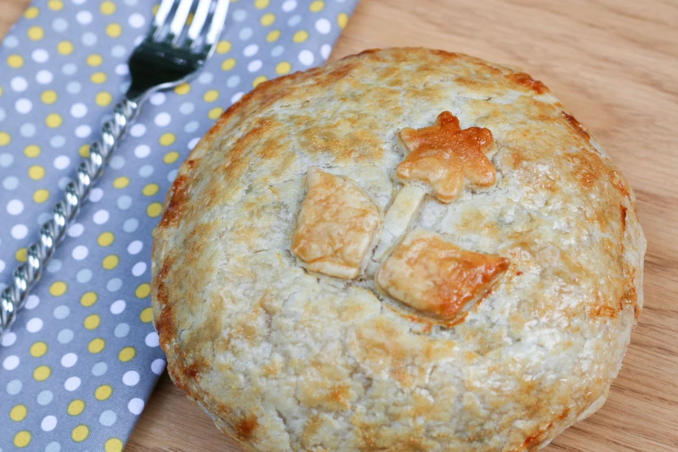 A round, golden-brown pie topped with pastry cutouts shaped like a flower and leaves, placed on a wooden surface beside a polka dot napkin and a fork.