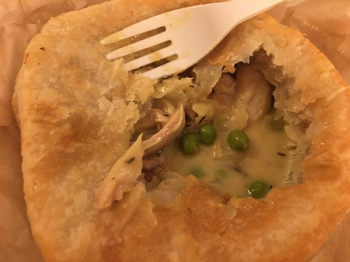 A close-up of a partially eaten pot pie with flaky golden crust, filled with creamy sauce, green peas, and pieces of chicken. A white plastic fork rests on top of the pie.