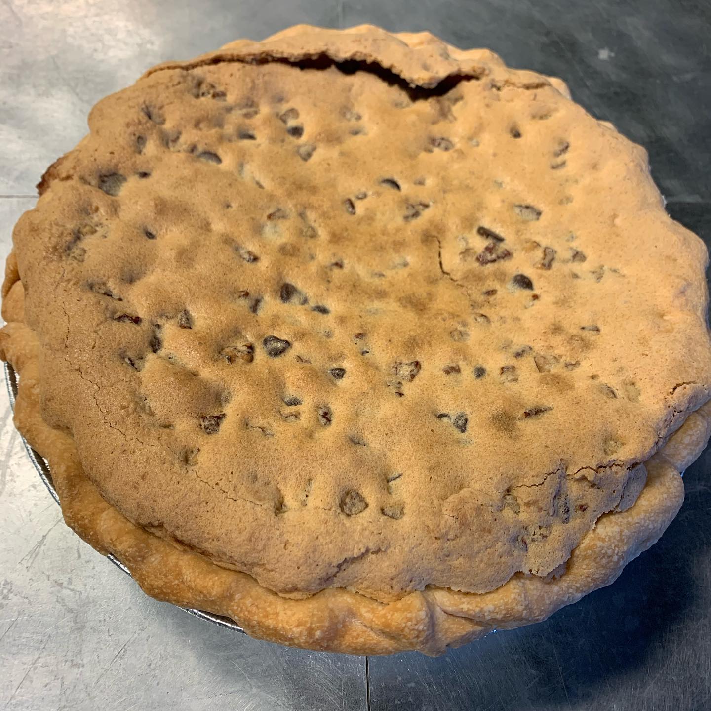 A large, round chocolate chip cookie pie with a golden-brown crust sits on a metal surface. The pie is thick and dotted with chocolate chips throughout the top.
