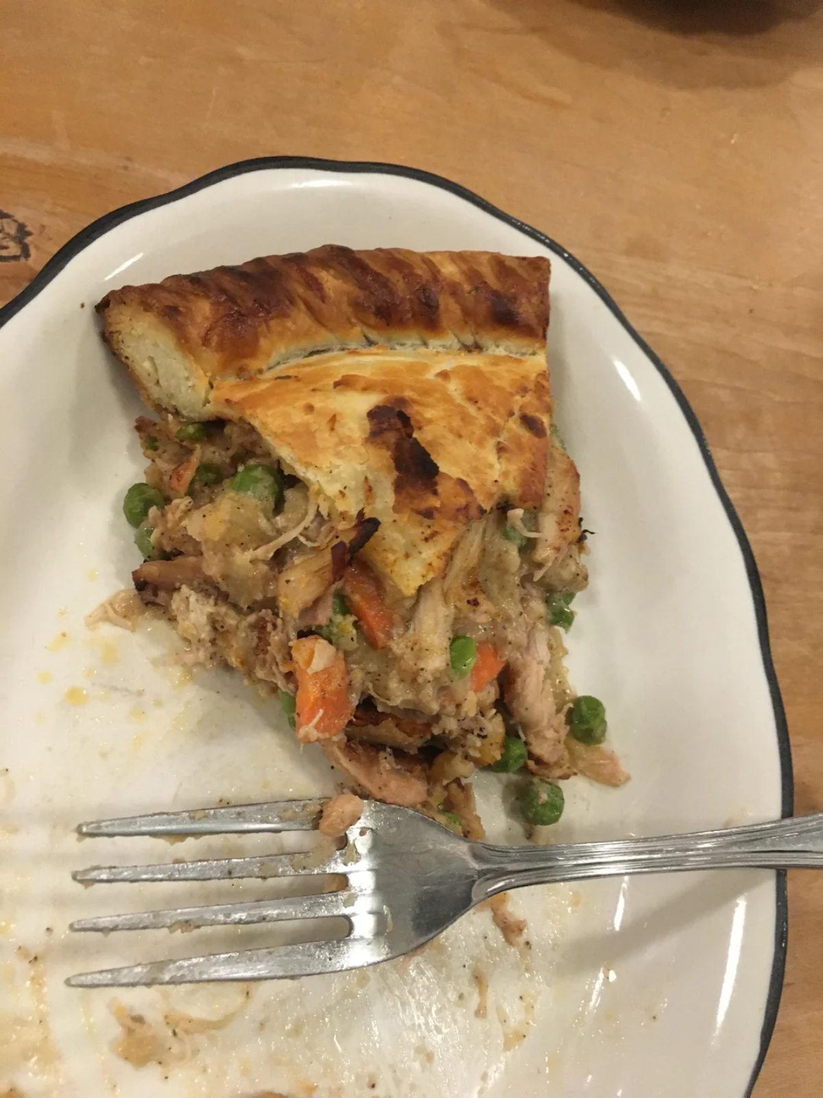 A partially eaten slice of chicken pot pie with vegetables on a white plate with a silver fork, set on a wooden table.