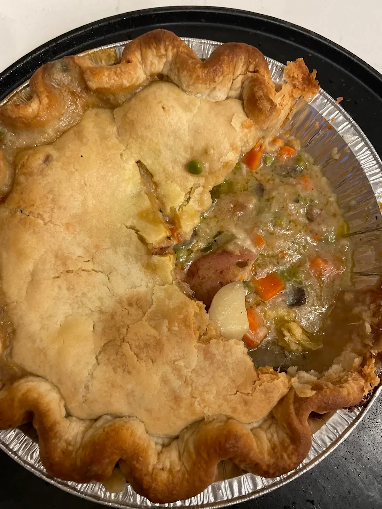 A partially eaten pot pie in a foil pie pan, showing a golden, flaky crust and a filling of vegetables and sauce, including potatoes, carrots, peas, and other ingredients.