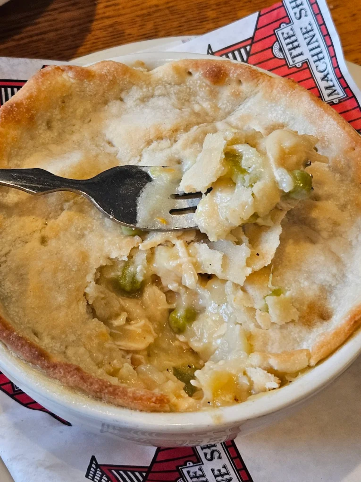 A fork lifting a piece of flaky crust from a bowl of chicken pot pie, showing creamy filling with peas and chicken inside, on a plate lined with a red and white napkin.