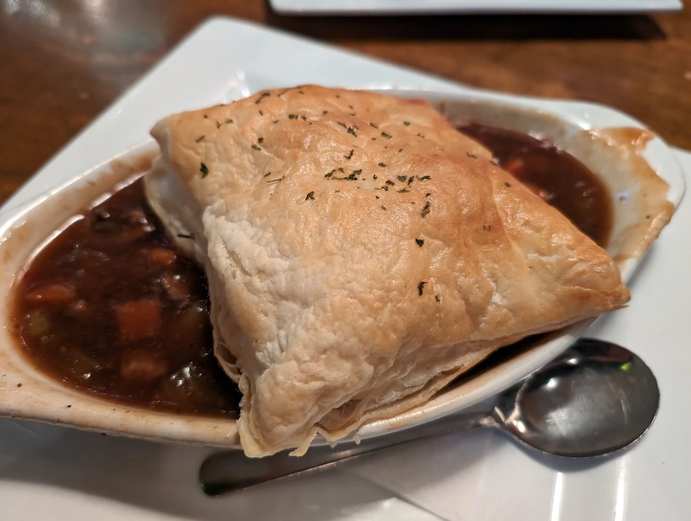 A close-up of a bowl of meat and vegetable stew topped with a flaky, golden puff pastry crust, served on a white plate with a spoon beside it.