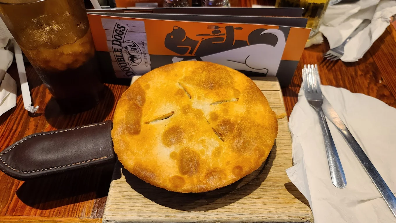 A golden-brown pot pie sits on a wooden board with a leather-handled tray. A drink, utensils, and a Double Dogs restaurant menu featuring a dog illustration are visible in the background.