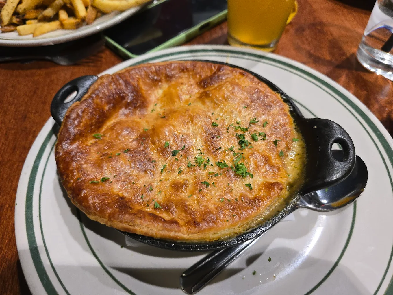 A golden-brown pot pie topped with herbs sits in a black baking dish on a white plate, accompanied by a spoon and fork. Other food and drinks are partially visible in the background on a wooden table.