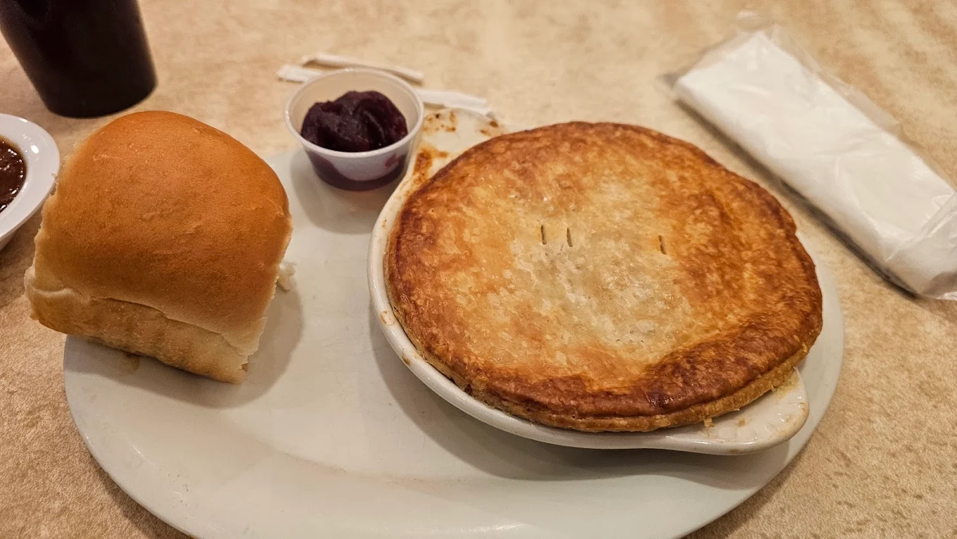 A round pot pie with a golden crust sits on a white plate next to a soft dinner roll and a small cup of grape jelly, with a napkin and utensils in the background on a beige table.