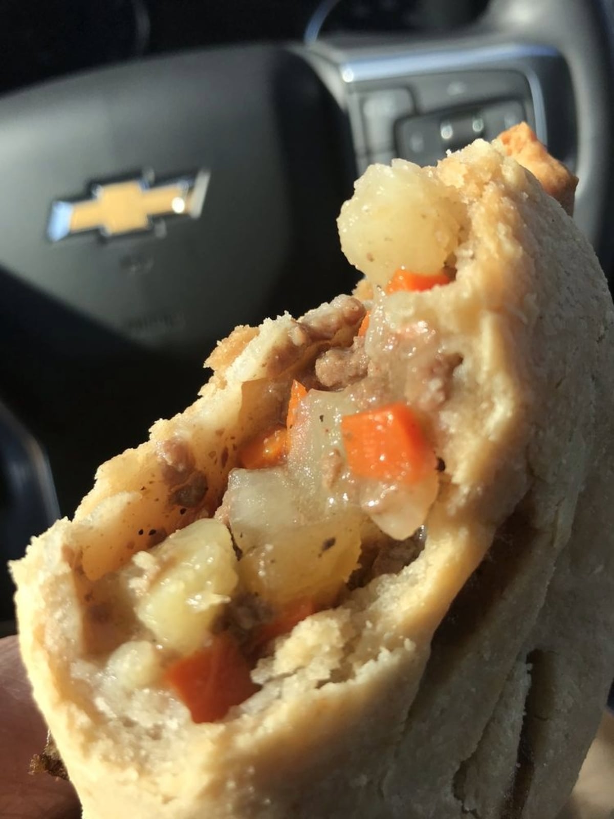 A close-up of a partially eaten pasty filled with diced potatoes, carrots, and meat, held in front of a car steering wheel with a Chevrolet logo.