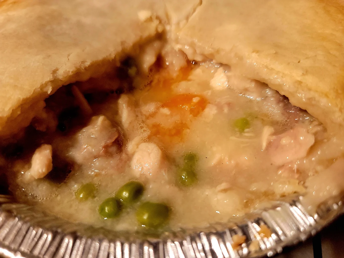 Close-up of a chicken pot pie in a foil tin with a portion cut out, revealing creamy filling with chicken pieces, peas, carrots, and sauce under a golden-brown crust.