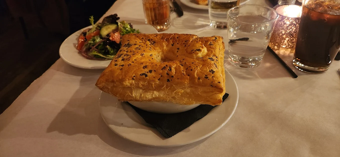 A bowl covered with golden, flaky puff pastry on a plate with a black napkin, next to a small salad, glasses of water, and iced drinks on a white tablecloth.