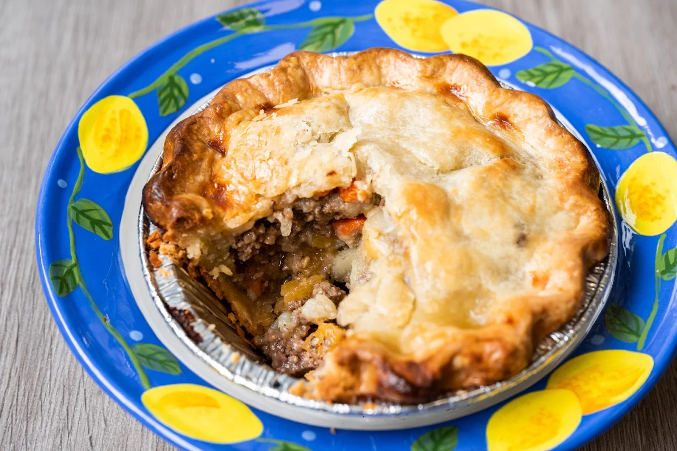 A savory meat pie with a golden, flaky crust sits on a blue plate decorated with yellow lemons. A slice of the pie has been removed, revealing a filling of ground meat and vegetables.