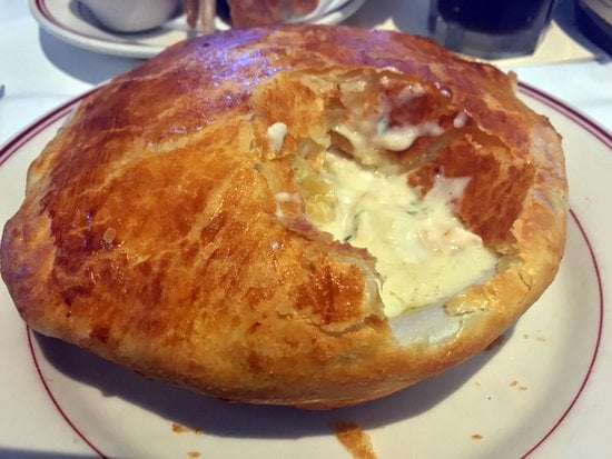 A golden-brown, flaky bread or pastry crust covers a creamy, white filling, served on a white plate with a red rim. Part of the crust has been broken, exposing the filling inside.