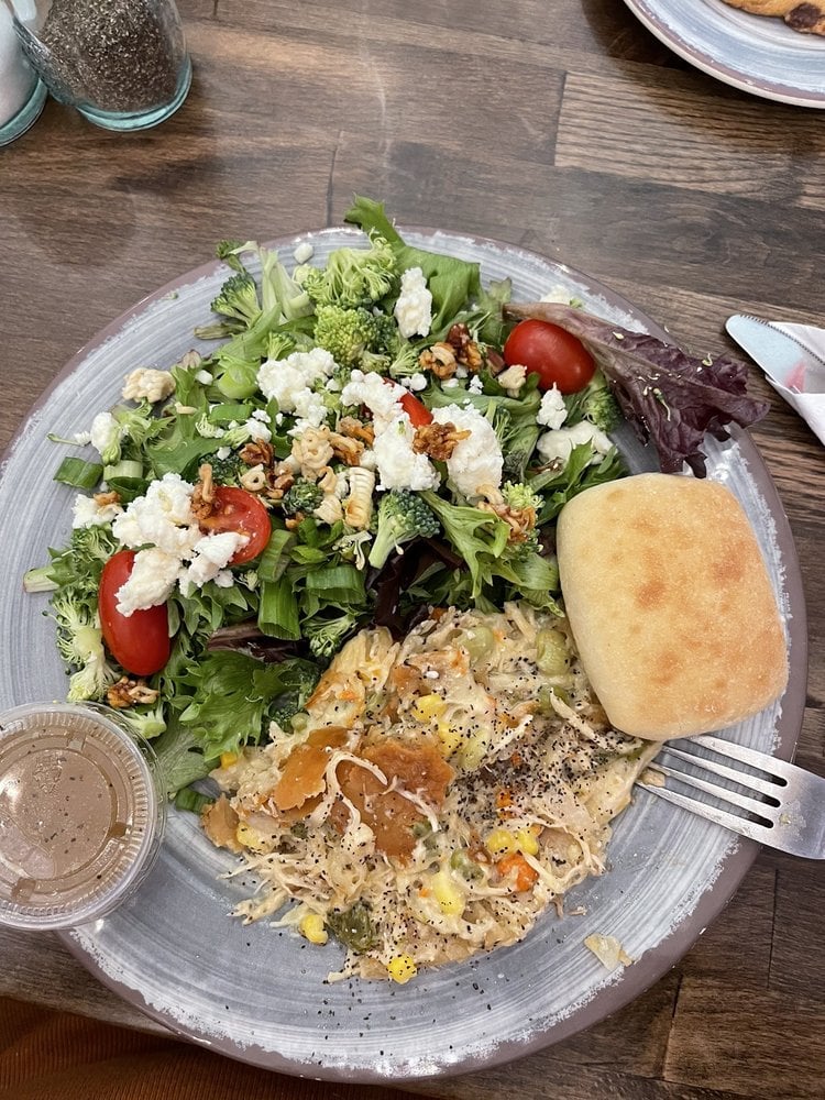 A plate with a green salad topped with cherry tomatoes, broccoli, walnuts, feta cheese, a bread roll, a serving of creamy chicken casserole, and a small container of salad dressing on the side.