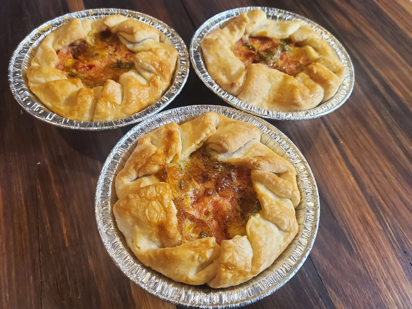 Three small savory pies with golden, flaky crusts are in individual aluminum tins on a dark wooden surface. The pies appear homemade, with visible herbs and a slightly rustic appearance.