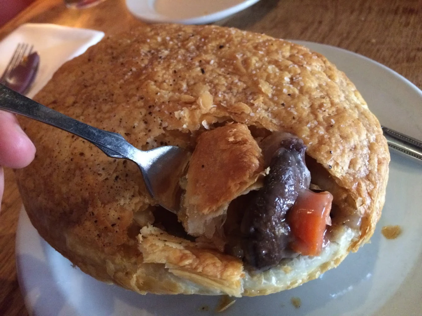 A fork breaks into a golden, flaky meat pie on a white plate, revealing chunks of beef, carrots, and gravy inside. The pie sits on a wooden table with a napkin and utensils in the background.