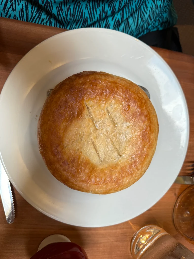 A golden-brown pot pie with a flaky crust sits on a white plate. Three short slits are cut into the top crust. Part of a knife, fork, glass, and a person wearing a blue patterned shirt are visible around the plate.