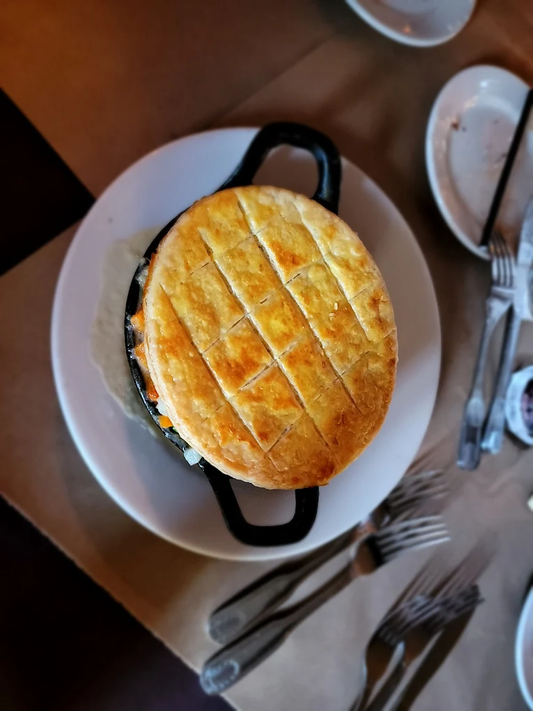 A round cast iron dish topped with a golden, lattice-patterned puff pastry sits on a white plate, surrounded by empty plates, forks, and knives on a brown paper-covered table.