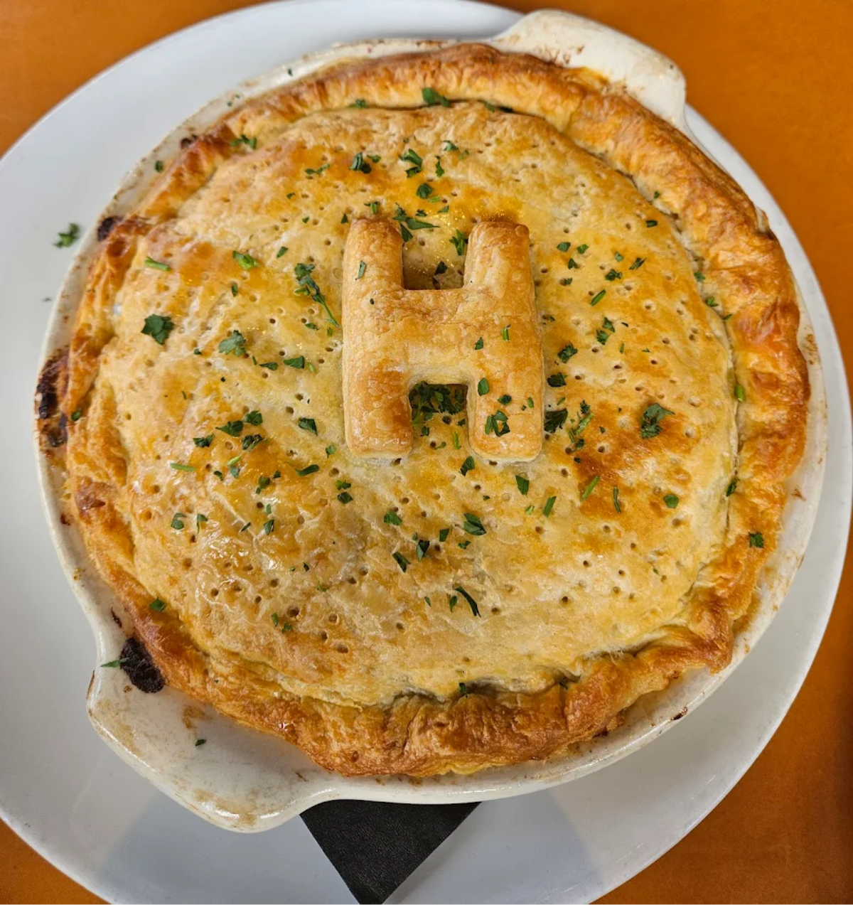 A golden-brown pot pie with a flaky crust sits on a white plate. The crust is topped with a pastry shaped like the letter "H" and sprinkled with chopped herbs.